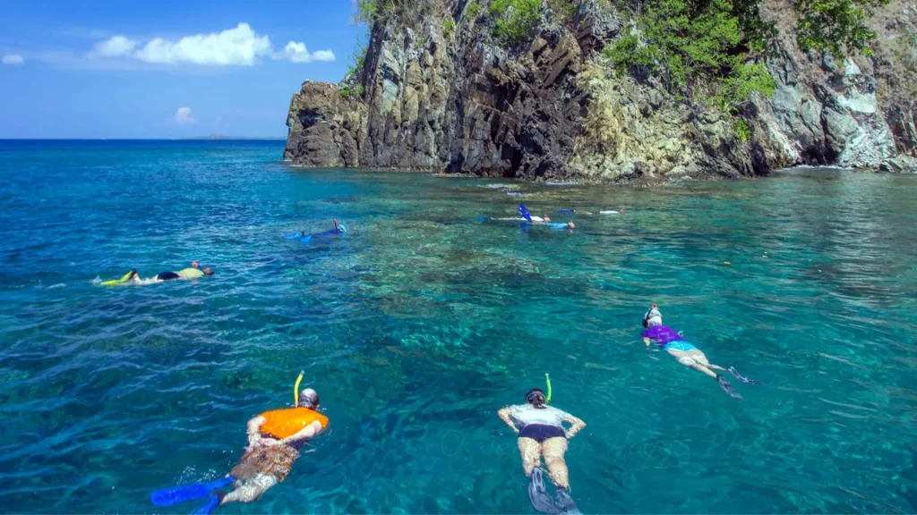 Scenic panoramic view of Crystal Bay Nusa Penida featuring crystal-clear turquoise waters with 30-meter visibility, soft white sand beach surrounded by lush green hills, and a small rocky islet offshore, accessible via Axe Stone Fast Cruise from Sanur Harbour to West Nusa Penida Tour.