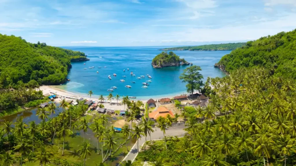 Spectacular view over Crystal Bay Nusa Penida with calm waters reflecting orange and pink hues, surrounded by coconut trees and coastal cliffs, one of the best sunset viewpoints in Nusa Penida accessible via Axe Stone Fast Cruise daily departures from Sanur Harbour.