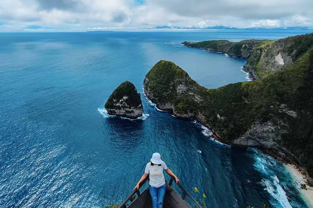Woman in white clothes sitting and capturing memories at Paluang Cliff on a West Nusa Penida Tour with Axestone Fast Cruise, overlooking the stunning ocean views. find this experiences on Nusa Penida Combo Tour